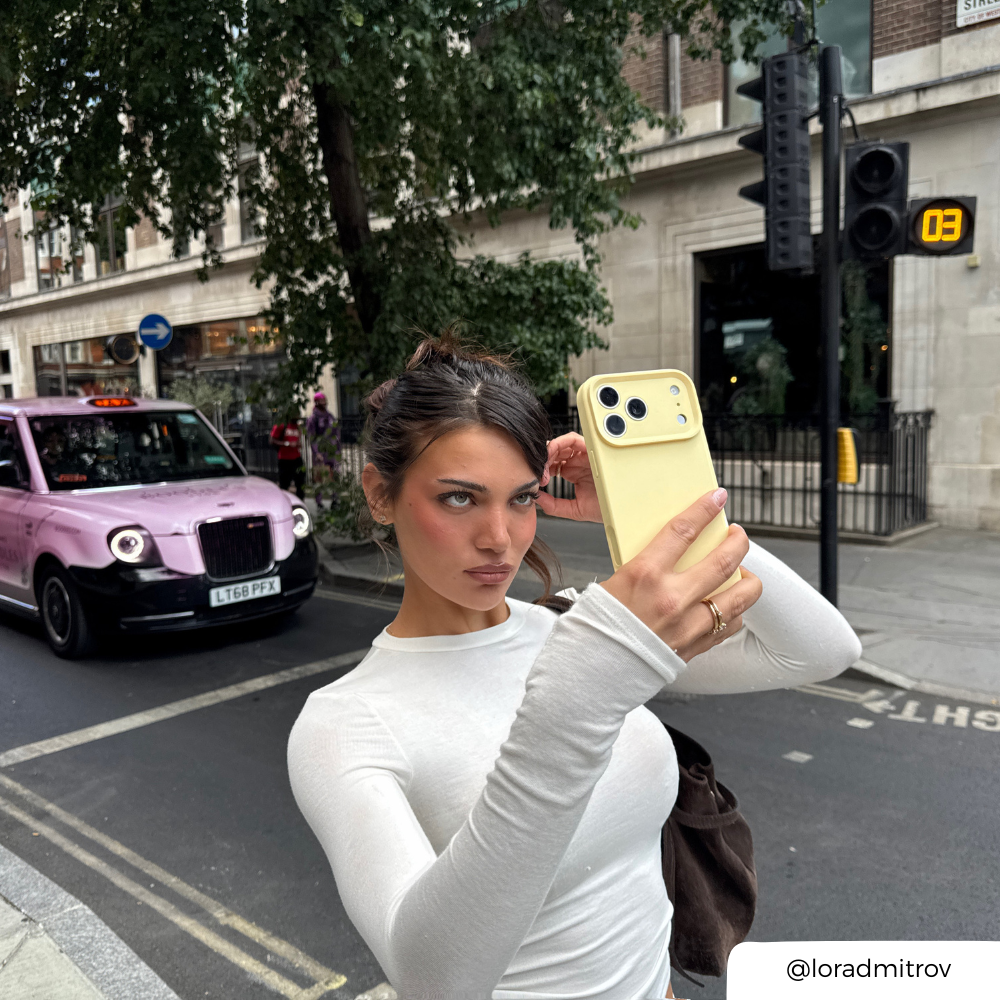 a woman taking a selfie in a street setting, holding an iphone in a yellow pastel phone case.