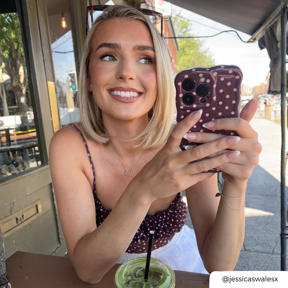 Woman holding an iphone with a brown with light pink polka dots phone case.