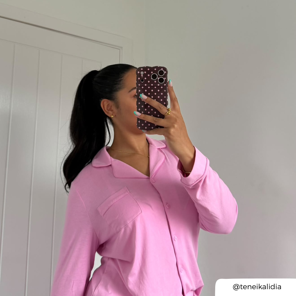 A woman takes a mirror selfie wearing a light pink long-sleeve button-up pyjamas. She is holding a dark brown polka-dot phone case in front of her face with baby pink spots.