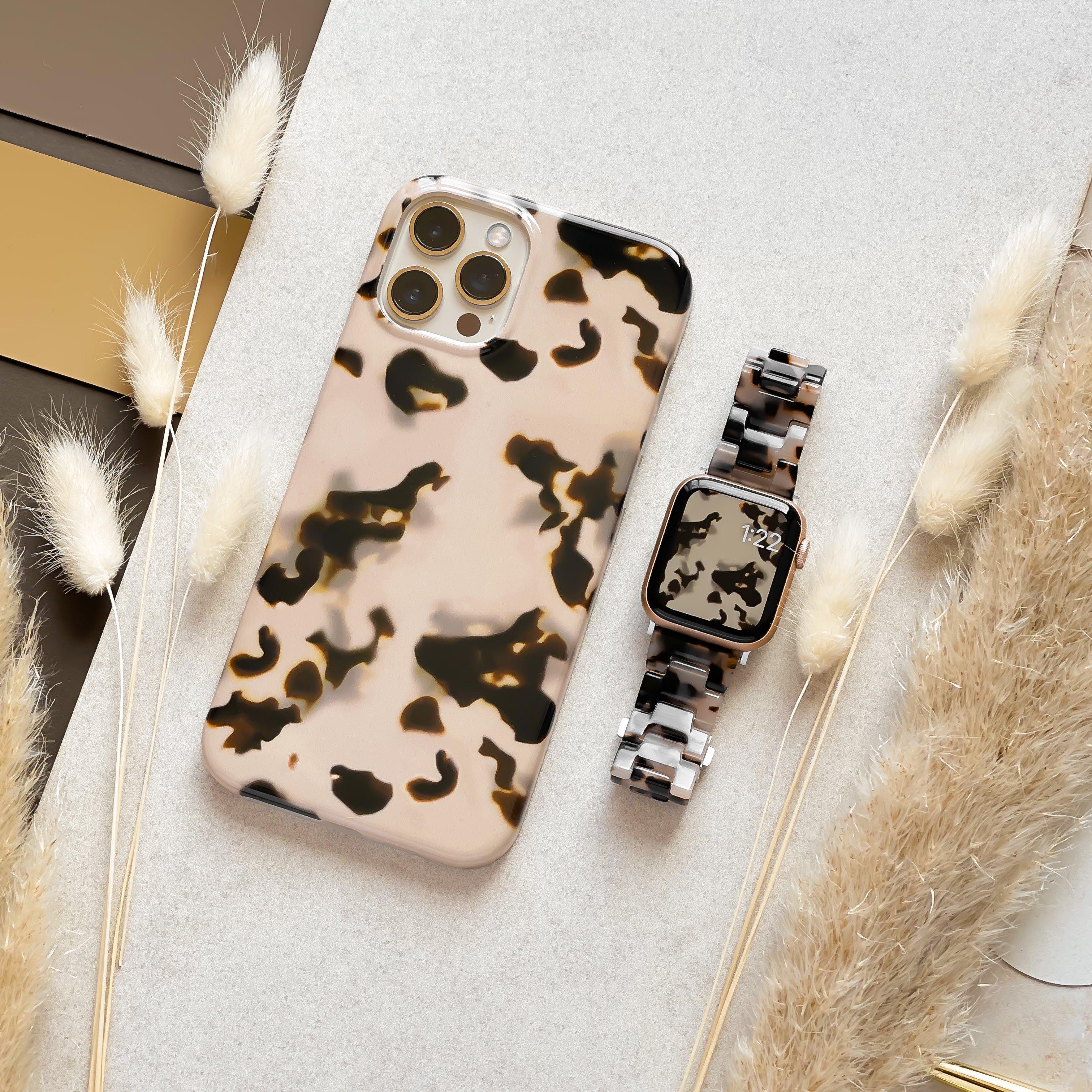 A smartphone in a beige camouflage case and a smartwatch featuring the Luxe Ivory Tort Apple Watch Strap by Coconut Lane lie on a textured surface. Soft, fluffy stems and a reflective, striped background form the surroundings, creating a stylish and coordinated aesthetic.