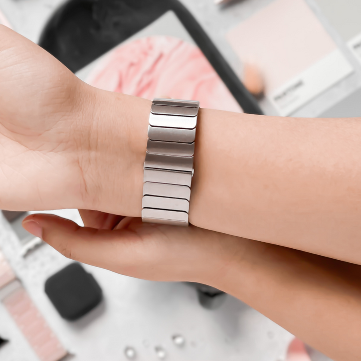 A close-up of a person adjusting a sleek Coconut Lane Slinky Apple Watch Strap in Silver on their wrist. The background features various pink and grey swatches, creating a stylish and aesthetically pleasing scene. An adjustable strap ensures the perfect fit, with a FREE link remover tool for easy customization.