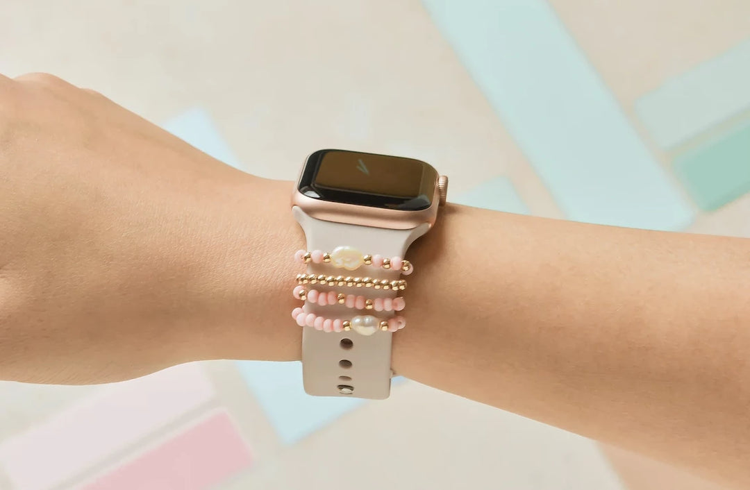 A person wearing a rose gold smartwatch with a white strap on their wrist. The wrist is adorned with pink and gold beaded bracelets. The background is softly blurred with pastel colors.