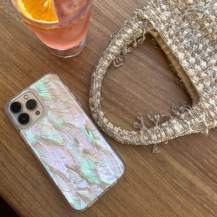 A Real Natural Shell Phone Case - White by Coconut Lane rests on a wooden table beside a textured handbag adorned with intricate beadwork. Nearby, there's a glass holding a beverage garnished with an orange slice.