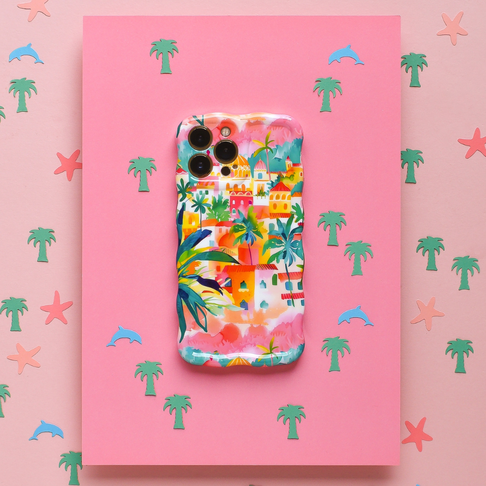 The Wavy Phone Case - Portofino Dreams by Coconut Lane is an extra protective phone case adorned with a vibrant tropical village design, featuring palm trees and houses. Set against a pink background dotted with small pastel palm tree, dolphin, and starfish icons, this case captures the coastal charm inspired by an Italian seaside getaway.