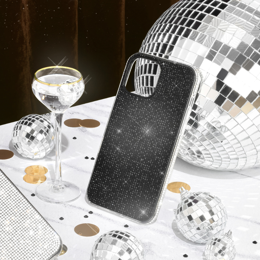 A Coconut Lane Disco Sparkle Phone Case - Black, adorned with shimmering rhinestones, is placed on a white table decorated with small and large disco balls. A clear champagne flute with a gold rim holds a sparkling beverage, and gold and black confetti enhance the festive ambiance.