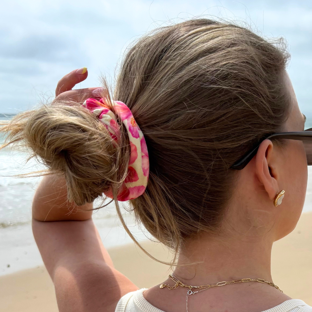 Woman in sunglasses wears a yellow scrunchie with hibiscus print by Coconut Lane, standing on a sandy beach.