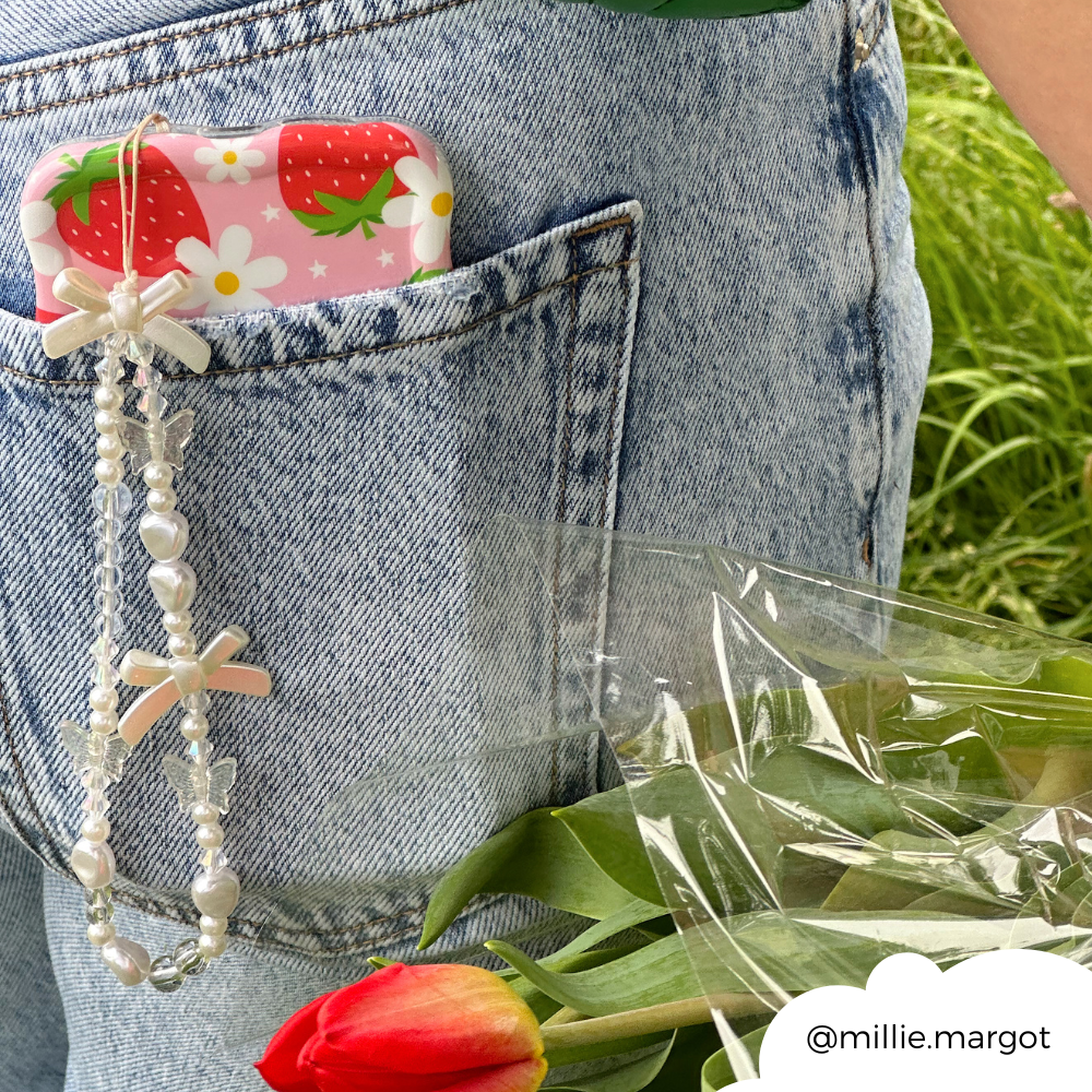 A close-up of a person wearing jeans with a phone case featuring strawberries and flowers peeking out of the pocket. Attached to the phone accessory is the Beaded Phone Strap - Bows & Butterflies by Coconut Lane. In their hand, they hold a bouquet wrapped in plastic, and the bottom red tulip is visible.
