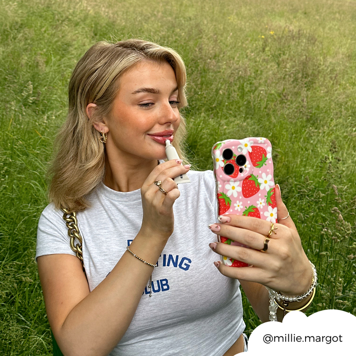 A woman with blonde hair, wearing a white "Fishing Club" t-shirt, applies lip gloss while taking a selfie with her phone, which is protected by the Wavy Phone Case - Berry Cute Strawberry from Coconut Lane. She is sitting in a grassy field. Her Instagram handle "@millie.margot" is visible in the bottom right corner.