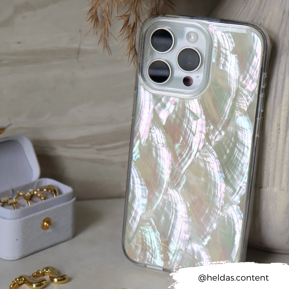 A silver iPhone with a gleaming Real Natural Shell Phone Case - White from Coconut Lane rests on a surface beside an open white box holding gold jewelry. The backdrop features dried foliage. The Instagram handle "@heldas.content" appears at the bottom right corner of the image.