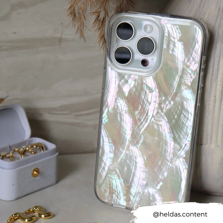 A silver iPhone with a gleaming Real Natural Shell Phone Case - White from Coconut Lane rests on a surface beside an open white box holding gold jewelry. The backdrop features dried foliage. The Instagram handle "@heldas.content" appears at the bottom right corner of the image.