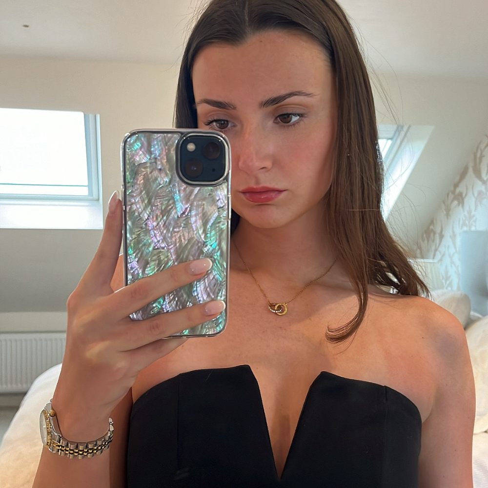 A young woman with long brown hair takes a mirror selfie inside a room with white walls and a window. She wears a strapless black top, a gold necklace with a crescent moon pendant, and a watch on her left wrist. Her phone case, the Real Natural Shell Phone Case in White by Coconut Lane, displays an iridescent, shell-like pattern.