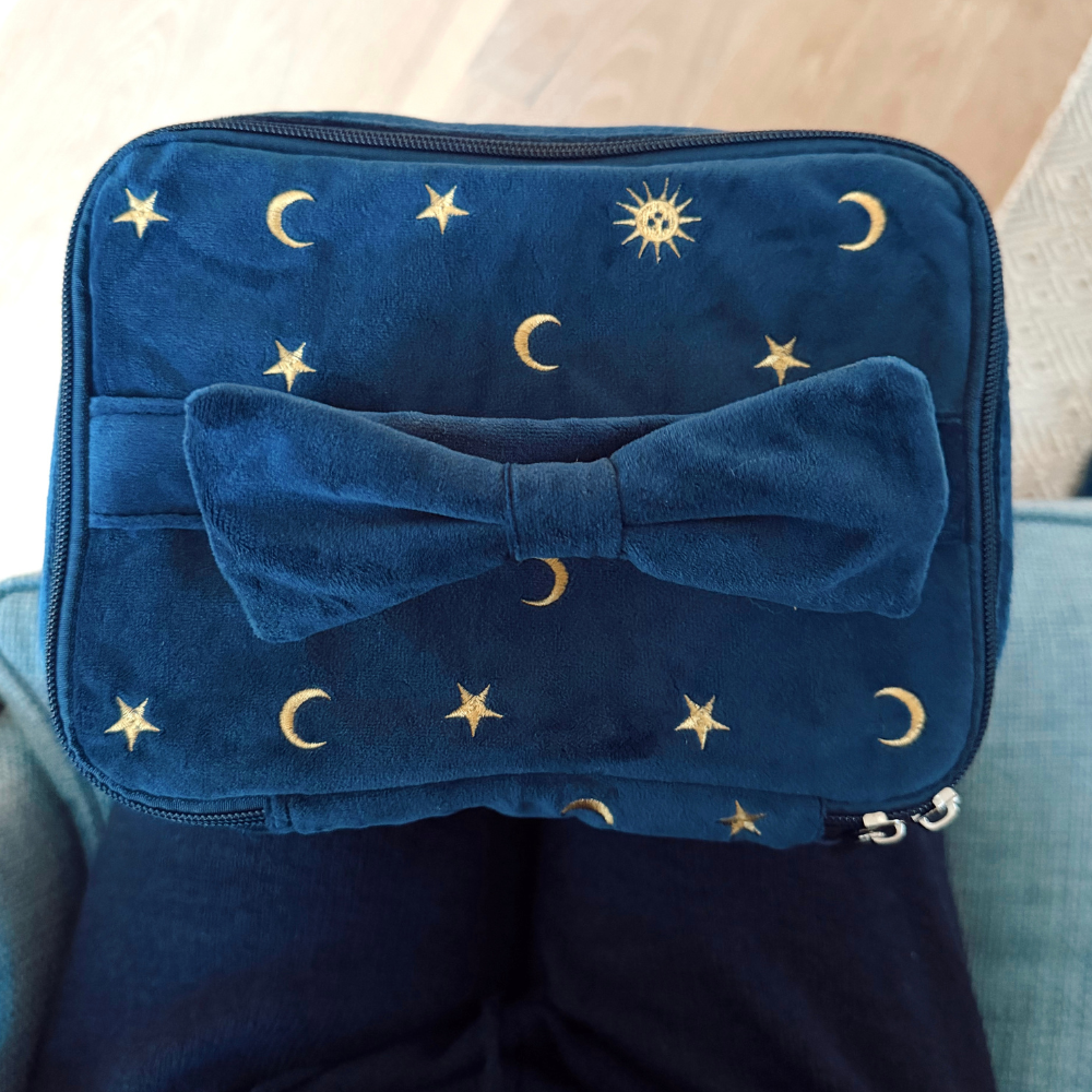 Blue velvet travel vanity case with a bow and gold celestial accents, shown resting on someones lap.