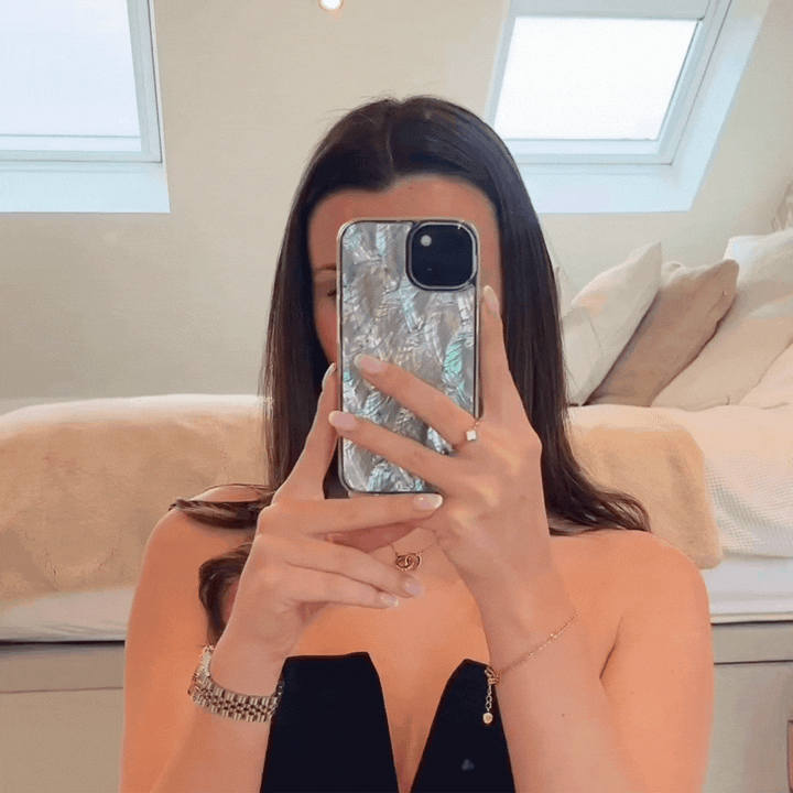 A woman with long dark hair is taking a mirror selfie using an iPhone protected by Coconut Lane’s Real Natural Shell Phone Case in White. She is dressed in a black top and minimal jewelry, including a bracelet and necklace. Behind her, you can see a neatly made bed and ceiling windows.