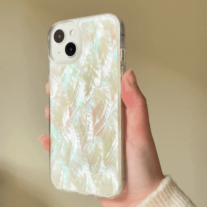 A hand holding an iPhone with Coconut Lane's Real Natural Shell Phone Case - White, which features a glossy, iridescent finish and a subtle feather-like pattern. The background is softly blurred. The case has a luminous, pearlescent sheen with hints of pink, blue, and green reflecting off its surface.