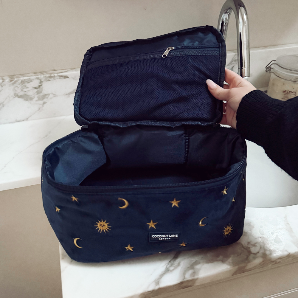 Hand opens Coconut Lane Travel Vanity Case in navy with gold star and moon pattern, next to a marble sink.