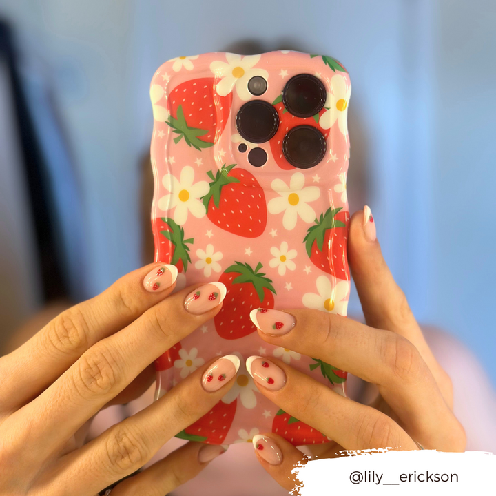 Close-up of hands holding an extra protective Coconut Lane Wavy Phone Case - Berry Cute Strawberry, featuring a pink design with strawberries and white flowers. The person's nails are painted white with red strawberry designs. Instagram handle @lily_erickson is in the bottom right corner.