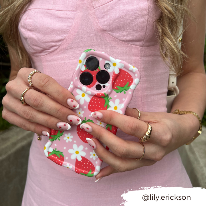 A person wearing a pink textured dress holds an extra protective Wavy Phone Case - Berry Cute Strawberry by Coconut Lane, adorned with strawberries and daisies. Their nails match the berry cute strawberry pattern. They are accessorized with multiple gold rings and a wristwatch. The text "@lily.erickson" appears at the bottom right.