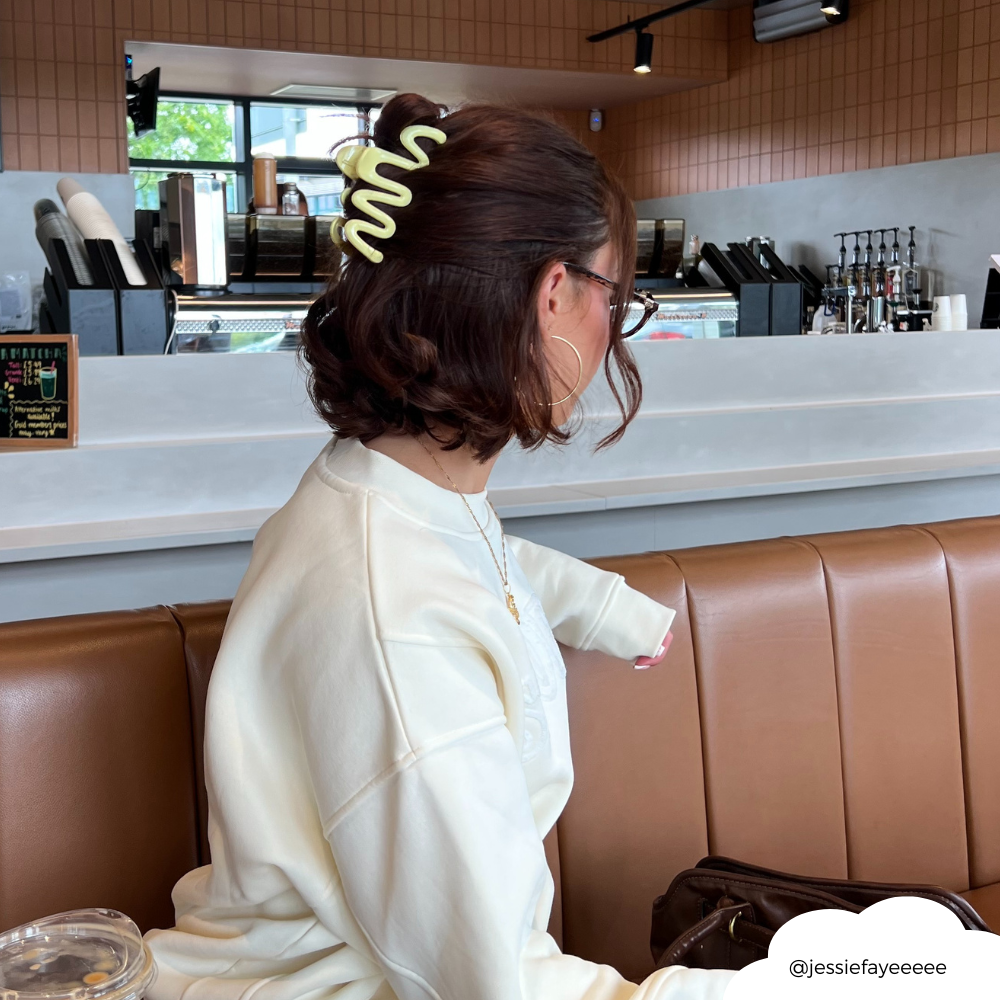 Yellow squiggle hair claw by Coconut Lane worn by a woman sitting on a tan booth in a modern café, seen from behind.
