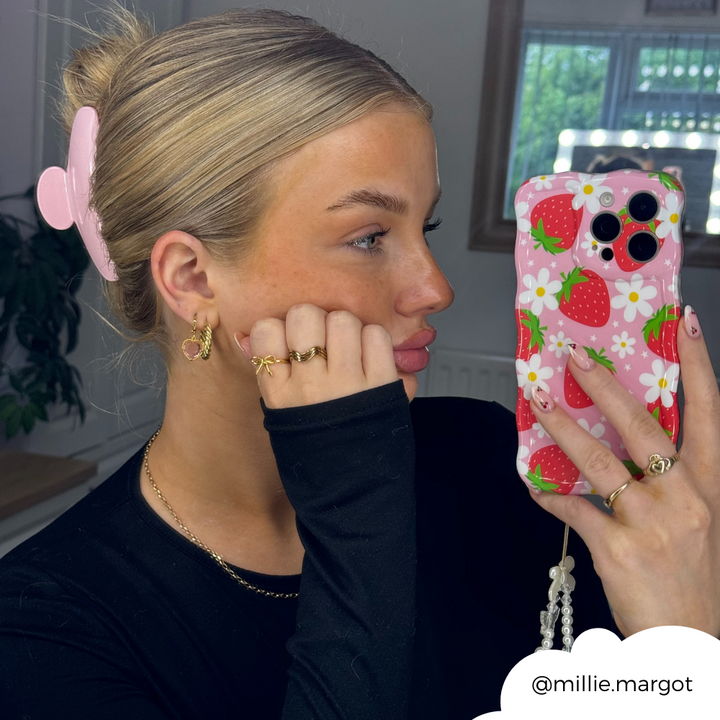 A person with blonde hair held back by a pink hair clip is taking a selfie in a mirror. They are wearing gold rings and earrings, a black top, and holding an extra protective phone encased in the Wavy Phone Case - Berry Cute Strawberry from Coconut Lane. The Instagram handle "@millie.margot" is shown in the image.