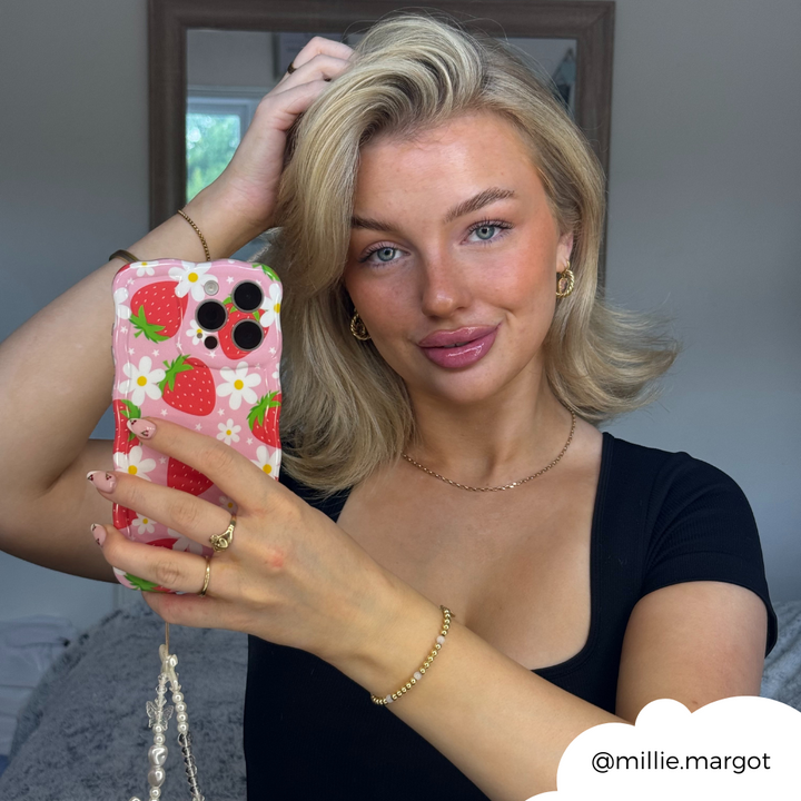A woman with shoulder-length blonde hair poses for a mirror selfie. She holds her Coconut Lane Wavy Phone Case - Berry Cute Strawberry. She wears gold jewelry and a black top. The Instagram handle "@millie.margot" is displayed in the bottom right corner.
