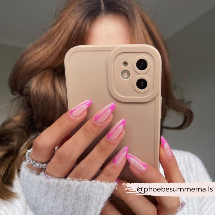 A person with wavy brown hair holds a beige phone, partially covering their face. Their long nails are manicured with a pink and white ombre design. They wear a white sweater and a ring with multiple stones. Text overlay says "@phoebesummernails". The Coconut Lane NAKD Phone Case - Nude offers full camera protection.