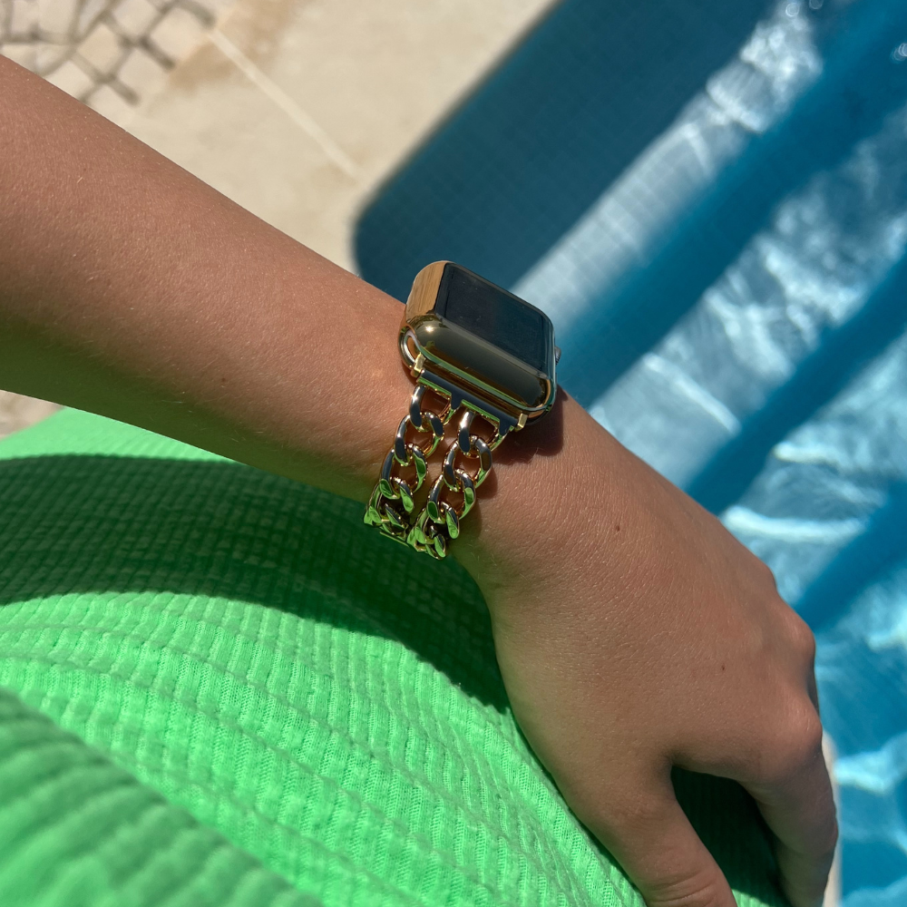 A person wearing a Dual-Chain Apple Watch Strap by Coconut Lane rests their hand on a green textured surface near a pool on a sunny day. The clear blue water of the pool is visible in the background.