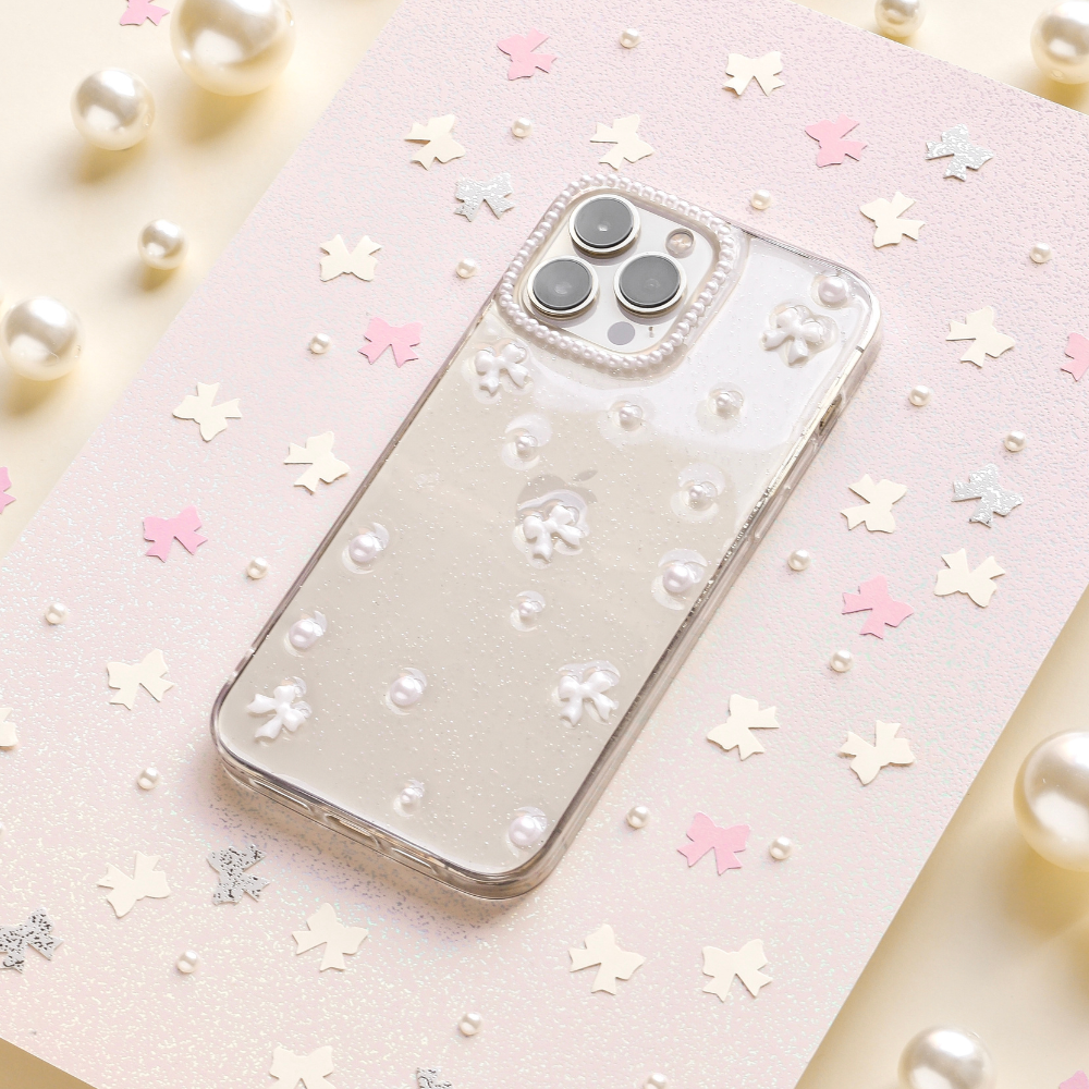 A smartphone adorned with the Mini White Bows Phone Case from Coconut Lane rests on a glittery pastel background, enhanced by scattered pink and silver star-shaped confetti and pearls. The phone’s triple camera setup is elegantly visible, contributing to a whimsical and sophisticated aesthetic.