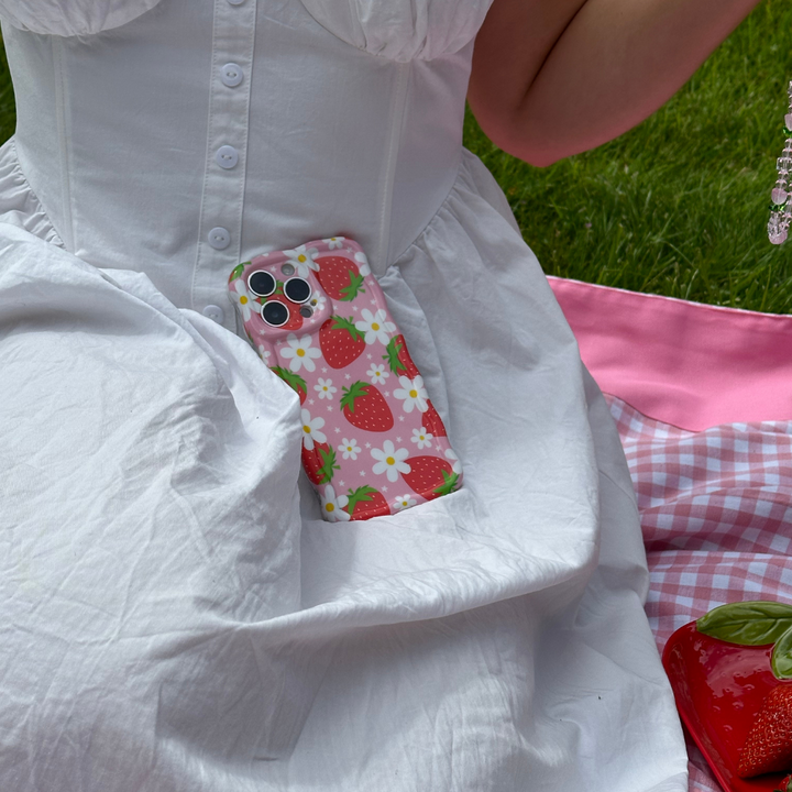 A person wearing a white dress sits on a pink and white checkered blanket on the grass. An extra protective smartphone with the Coconut Lane's Wavy Phone Case - Berry Cute Strawberry rests on their dress. Nearby, a red heart-shaped purse and a beaded accessory are visible.