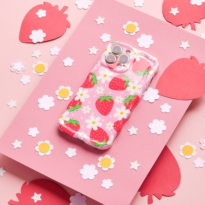 A Coconut Lane Wavy Phone Case - Berry Cute Strawberry with a delightful strawberry pattern and white flowers rests on a pink surface, surrounded by cut-out paper shapes of strawberries and flowers. The background includes larger pink strawberry shapes and smaller white flower shapes scattered around.