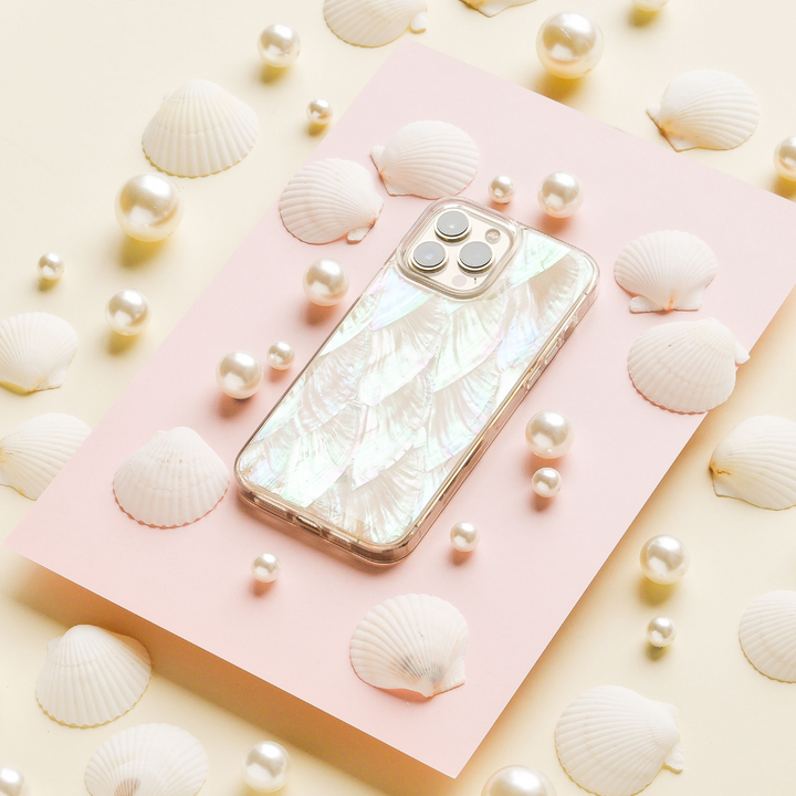 A smartphone with a pearlescent Real Natural Shell Phone Case - White by Coconut Lane is placed on a pink rectangular surface. The scene is decorated with assorted white seashells and scattered pearls, arranged on a cream-colored background.