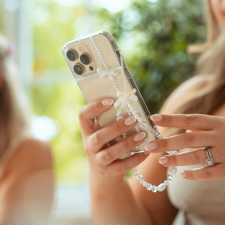 Close-up of a woman's hands holding an iPhone encased in a clear, beaded case adorned with small bows. The woman, who is wearing an engagement ring and Coconut Lane's Beaded Phone Strap - Bows & Butterflies, has manicured nails. The background features a soft blur of greenery and another person partially visible.