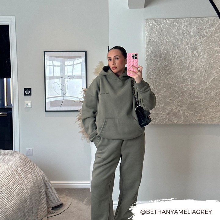 A woman is taking a mirror selfie in a modern, neutral-colored room, wearing an oversized olive-green hoodie and matching sweatpants. She's holding a candyfloss pink phone adorned with the Coconut Lane 3D Hearts Phone Case and carrying a black bag. The background features a framed abstract art piece and decorative pampas grass. Text: "@BETHANYAMELIAGREY.