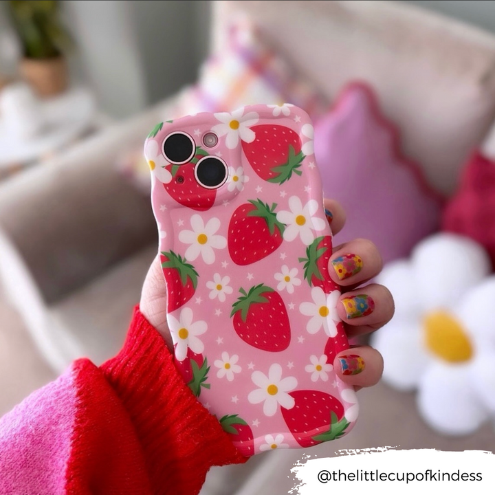 A hand with colorful, strawberry-themed nail art holds a phone encased in the extra-protective Coconut Lane Wavy Phone Case - Berry Cute Strawberry. The background showcases a blurred view of a cozy room with soft furnishings, including a pillow and a small table with a plant.