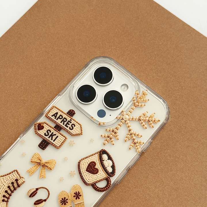 A clear phone case decorated with beige and brown ski-themed graphics, including skis, a pretzel, a coffee cup, a bow, and directional signs reading 'APRÈS' and 'SKI'.