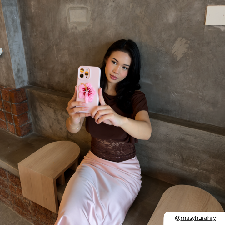 A woman takes a selfie while wearing a light pink satin skirt and a dark brown off-the-shoulder top, holding an iphone in a light baby pink phone case decorated with a beaded pink and red hibiscus flower phone patch. She is seated on a bench against a textured grey wall.