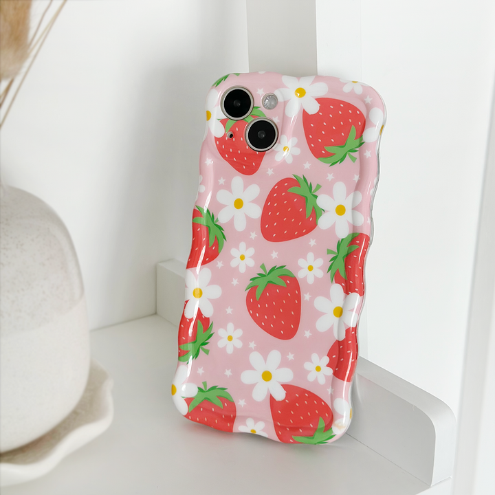 The Wavy Phone Case - Berry Cute Strawberry by Coconut Lane, adorned with red strawberries and white flowers, is propped against a white surface. A beige vase is partially visible in the background.