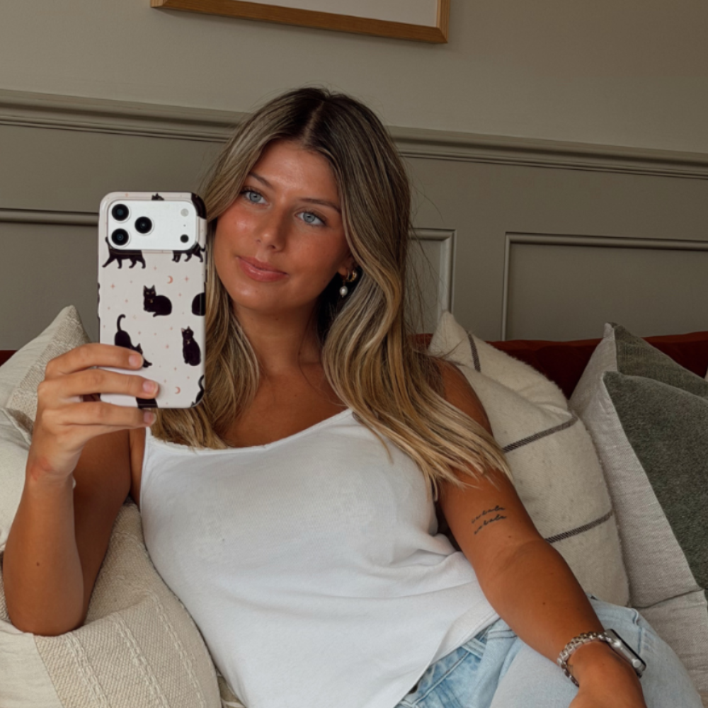 Woman holding up an iphone, showing a phone case with black cats printed on the case. She is sat on a sofa, wearing a white vest and blue jeans. 