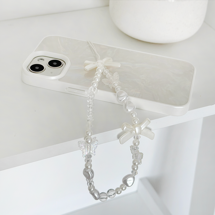 A white smartphone with a pearlescent case rests on a white surface. The Coconut Lane "Beaded Phone Strap - Bows & Butterflies," featuring beads, pearls, a butterfly, and a bow, adds an elegant touch as it dangles from the device. In the background, a white ceramic container is visible.