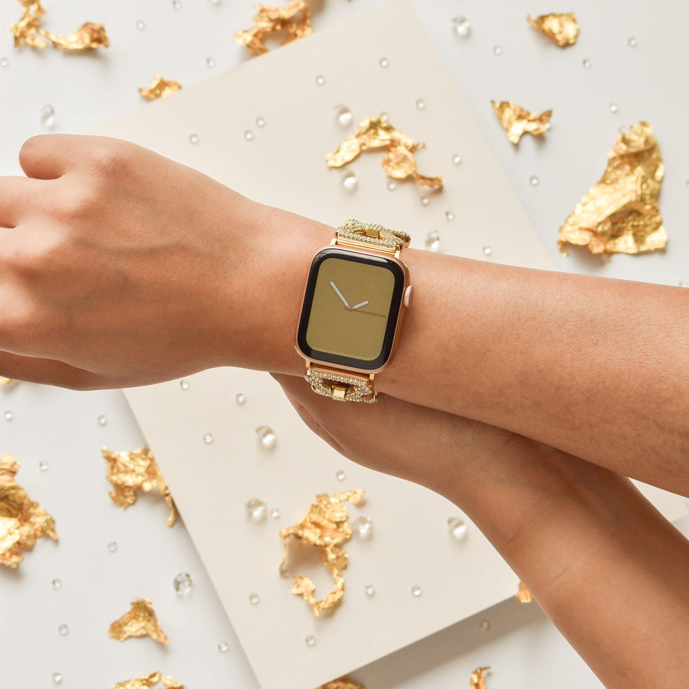 A wrist adorned with Coconut Lane’s Diamante Bracelet Apple Watch Strap, featuring a gold square face, set against a backdrop enlivened by gold foil fragments and scattered droplets. The strap, compatible with Apple Watch models, gleams as another hand lightly touches the wrist.