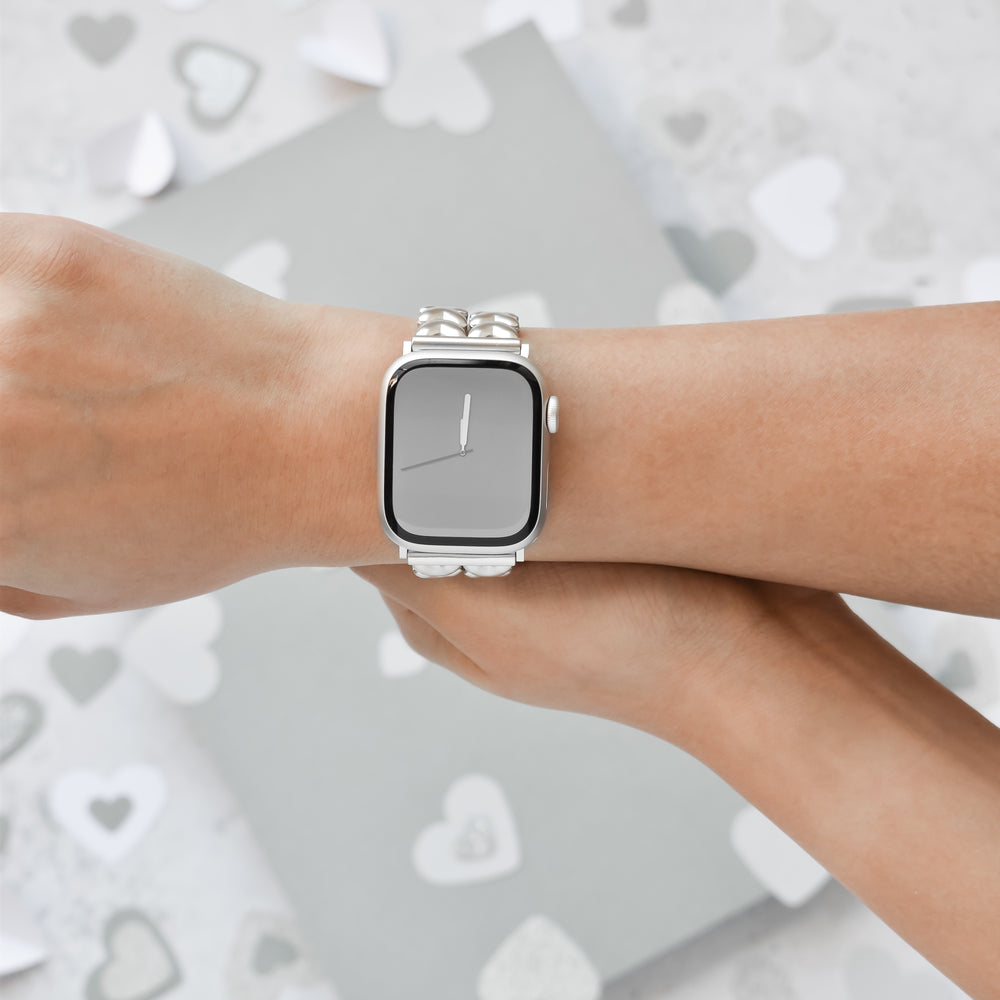 A person is wearing a smartwatch with a blank screen on their left wrist, featuring the Hearts Chain Apple Watch Strap – Starlight by Coconut Lane made of stainless steel. The background displays a blurred array of white and gray heart shapes. Their right hand lightly touches the left wrist, demonstrating how it fits ALL Apple Watch models.