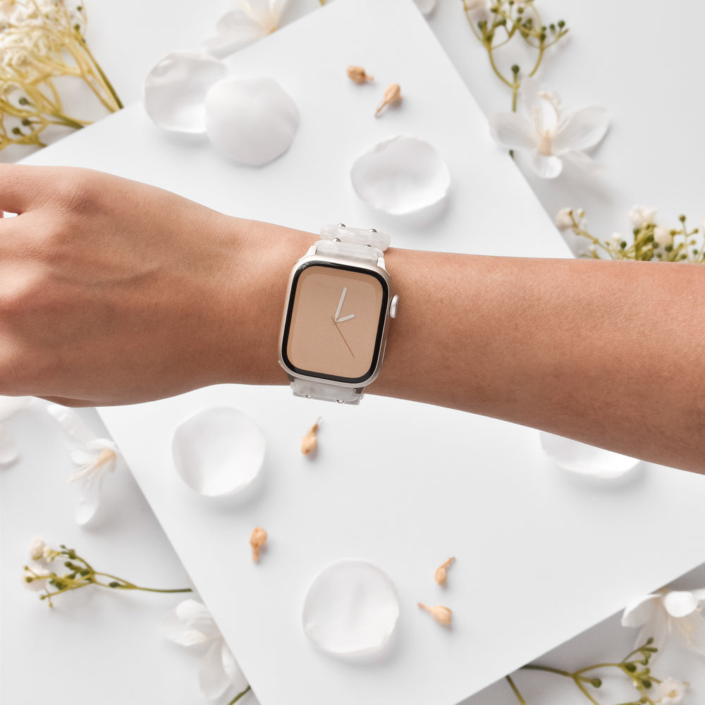 A left hand adorned with a stylish, minimalist watch featuring a beige face and no numbers. The timepiece is enhanced by Coconut Lane's unique White Pearl Beaded Apple Watch Strap. White flower petals and blossoms are scattered on the white surface in the background, creating a clean and elegant composition.