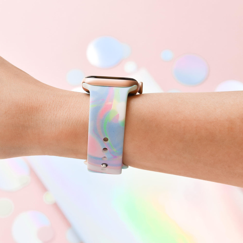 A close-up image of a person's arm wearing an Apple Watch paired with the Coconut Lane Holo Bubble Apple Watch Strap, showcasing its multi-colored, pastel gradient. The background is a soft pink with blurred circular reflections that create a dreamy, abstract atmosphere, perfectly accentuating the strap's holographic iridescent design.