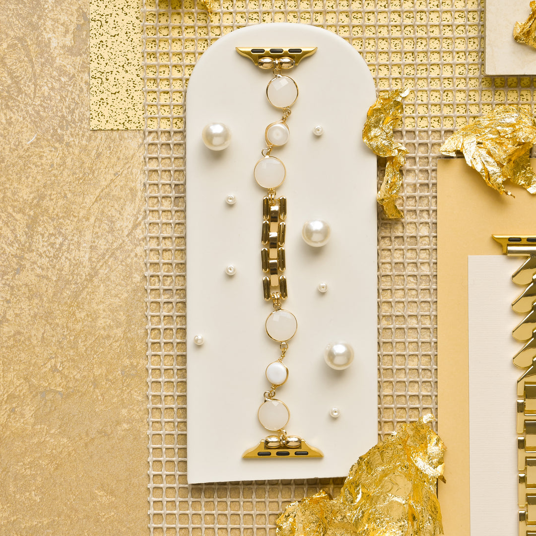 The Cute Stone Bracelet Apple Watch Strap by Coconut Lane is displayed on a white, arched stone-like background featuring golden and white textured decorative elements. This setup, which exudes the elegance comparable to an Apple Watch strap, rests on a gold metallic surface, creating an exquisitely luxurious look.