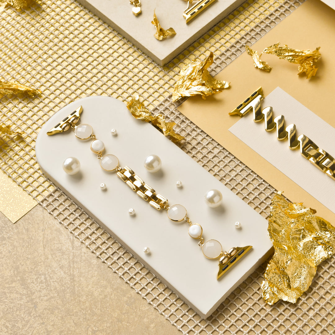 A stylish composition of gold-tone hair accessories and a Coconut Lane Cute Stone Bracelet Apple Watch Strap with pearl details displayed on a white, arched platform. Surrounding the accessories are golden leaf fragments and a textured gold background, creating an elegant and luxurious scene.