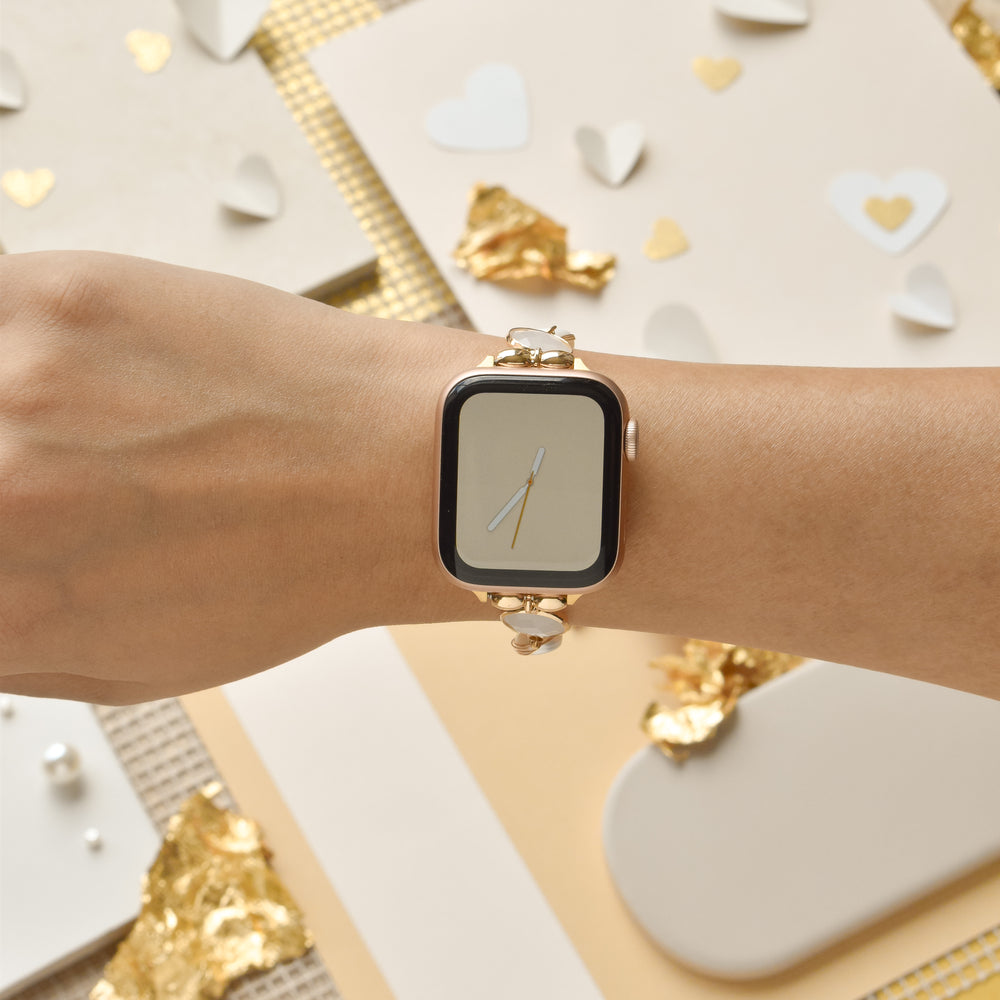 A person's wrist wearing a gold smartwatch with a simple clock face is shown, beautifully complemented by the Cute Stone Bracelet Apple Watch Strap from Coconut Lane. The background features various decorative items such as white paper hearts, gold foil pieces, and textured fabric, creating an elegant and festive atmosphere.