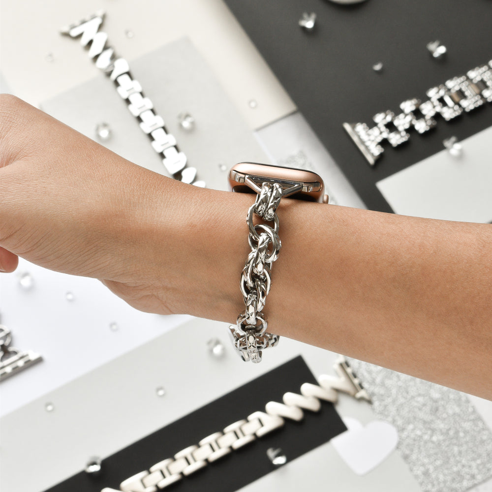 A person wearing the Coconut Lane Chain Bracelet Apple Watch Strap - Silver on their wrist. The background reveals various silver jewelry pieces and accessories displayed on different textured surfaces, including an elegant silver chain bracelet.