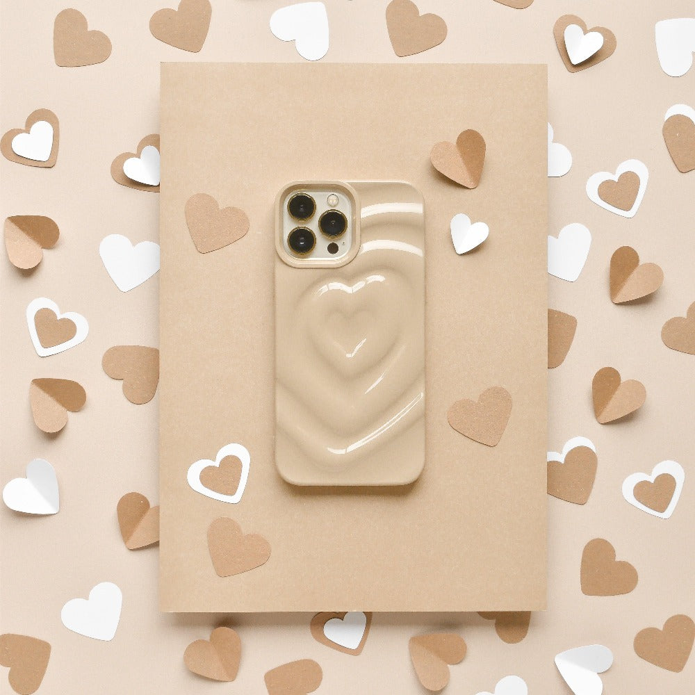 A "Melting Heart Phone Case - Biscuit Beige" from Coconut Lane, adorned with an extra protective cover featuring a raised 3D heart design, rests on a beige rectangular surface. The area around it is decorated with small white, beige, and brown paper hearts scattered evenly.