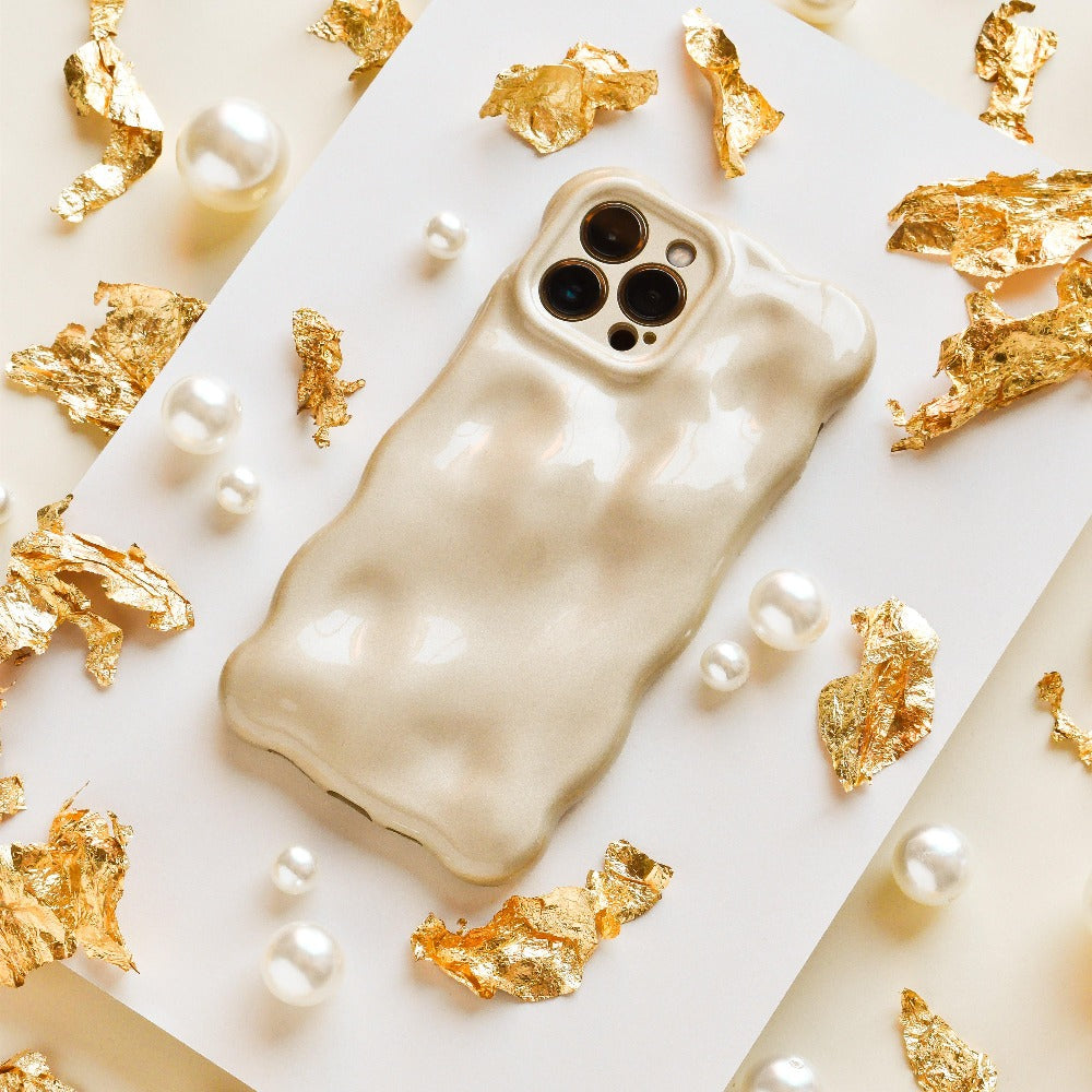 Displayed on a flat surface, surrounded by decorative gold foil fragments and scattered pearl beads, is the textured, glossy beige 3D Pearl Phone Case - Gold from Coconut Lane. This stylish and luxurious case features a wavy design and has precise cutouts for the camera lenses.