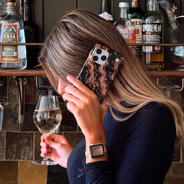 A woman with blonde-highlighted hair stands in a bar setting, holding a wine glass and covering part of her face with a iphone in a beige and dark brown marble design with subtle gold glitter flecks phone case