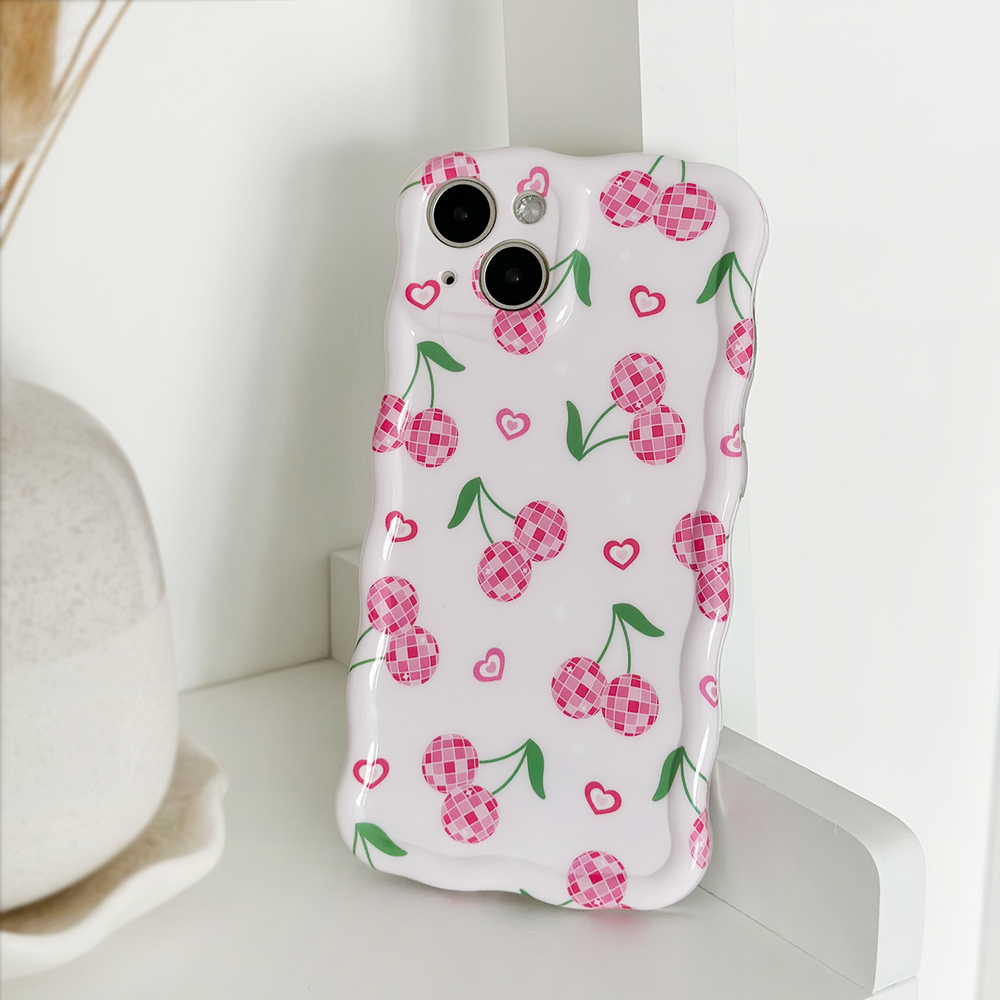 The Wavy Phone Case - Disco Cherries by Coconut Lane, featuring pink checkered cherries and small hearts on a white background, is propped up against a white surface. The glossy finish adds a touch of elegance, while in the background, part of a vase on a plate is visible.